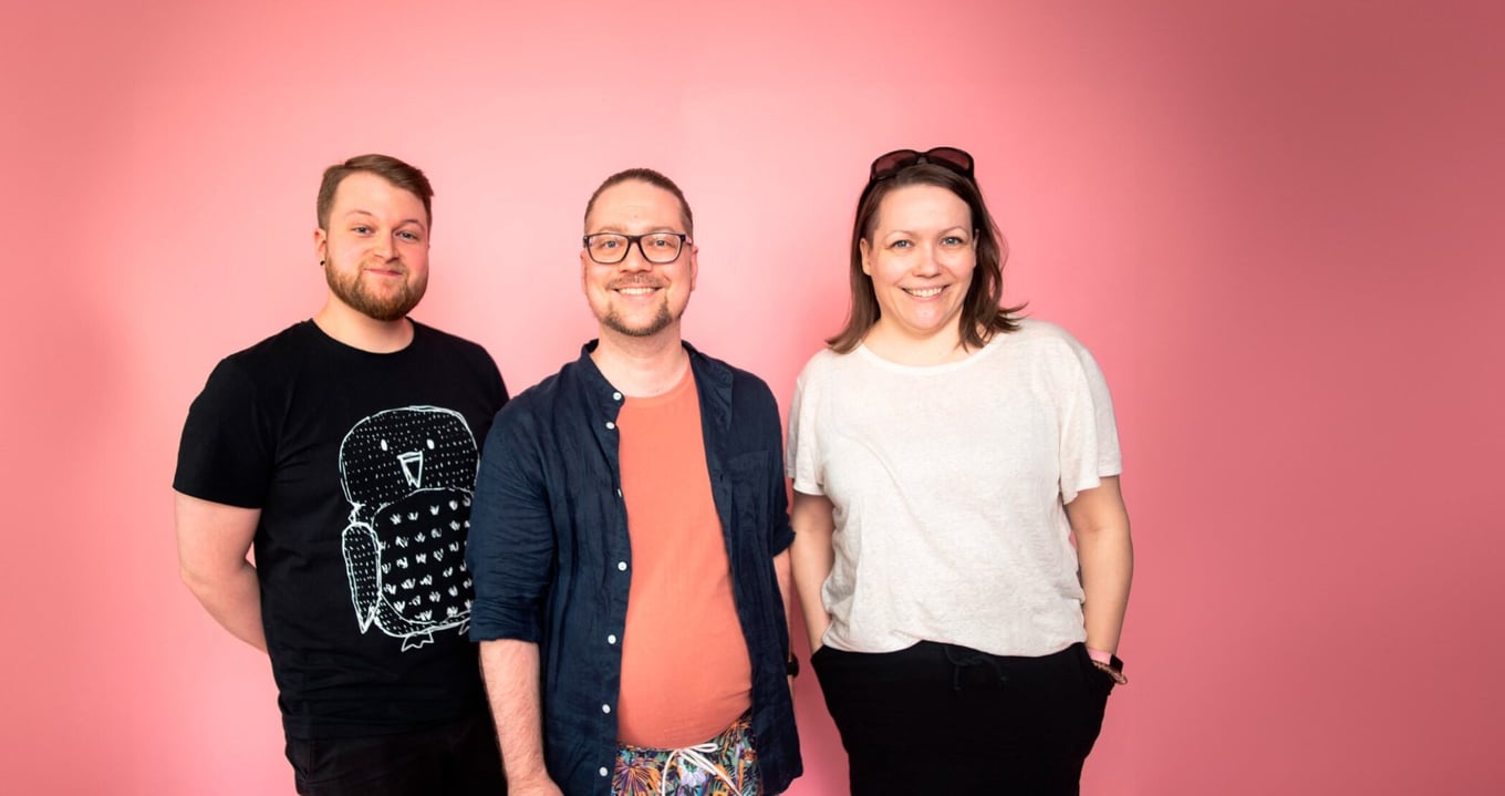 Three fraktio's workers stand in a row smiling in front of a pink background