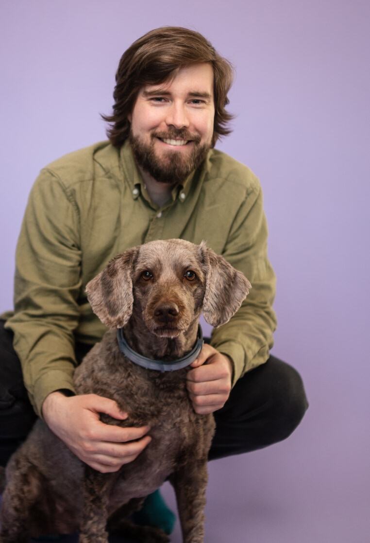 Developer Tatu is crouching in front of the purple background together with his dog Myrsky.